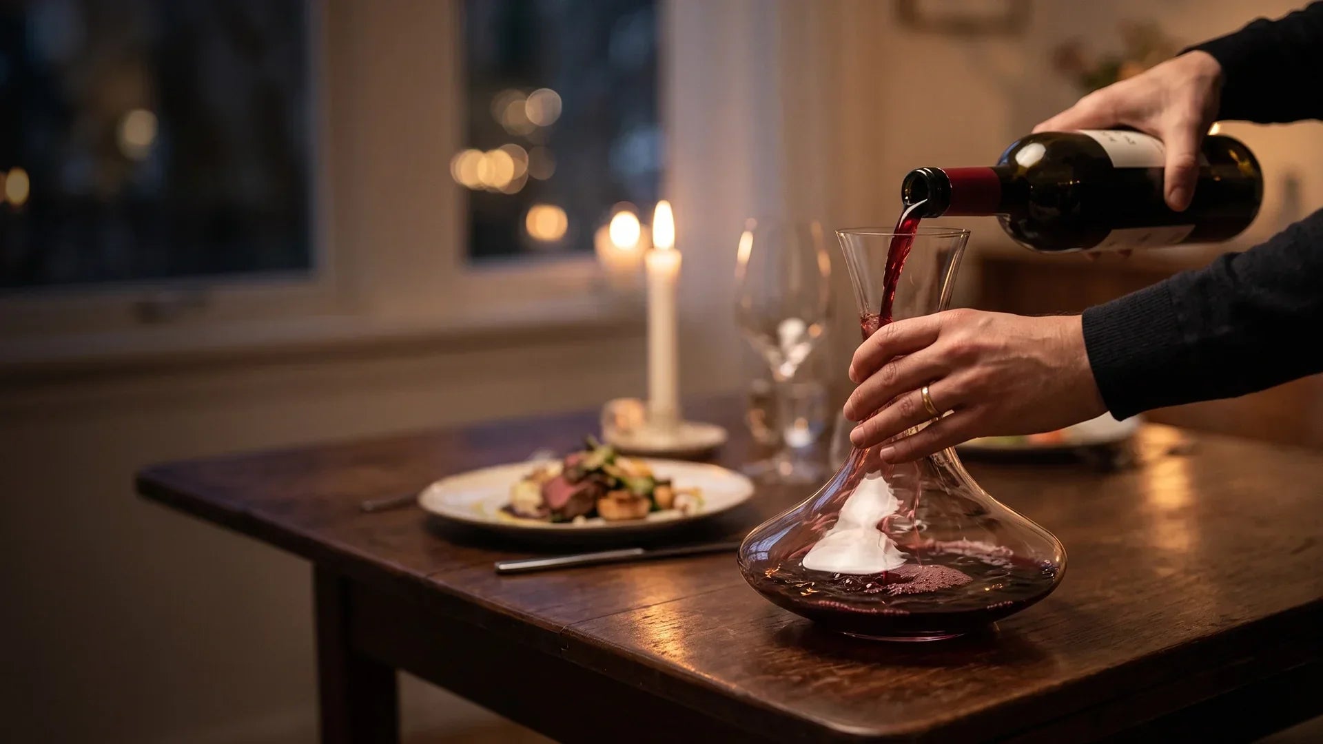 Red wine being poured into a wine decanter with a built-in aerating chamber on a rustic wooden table.