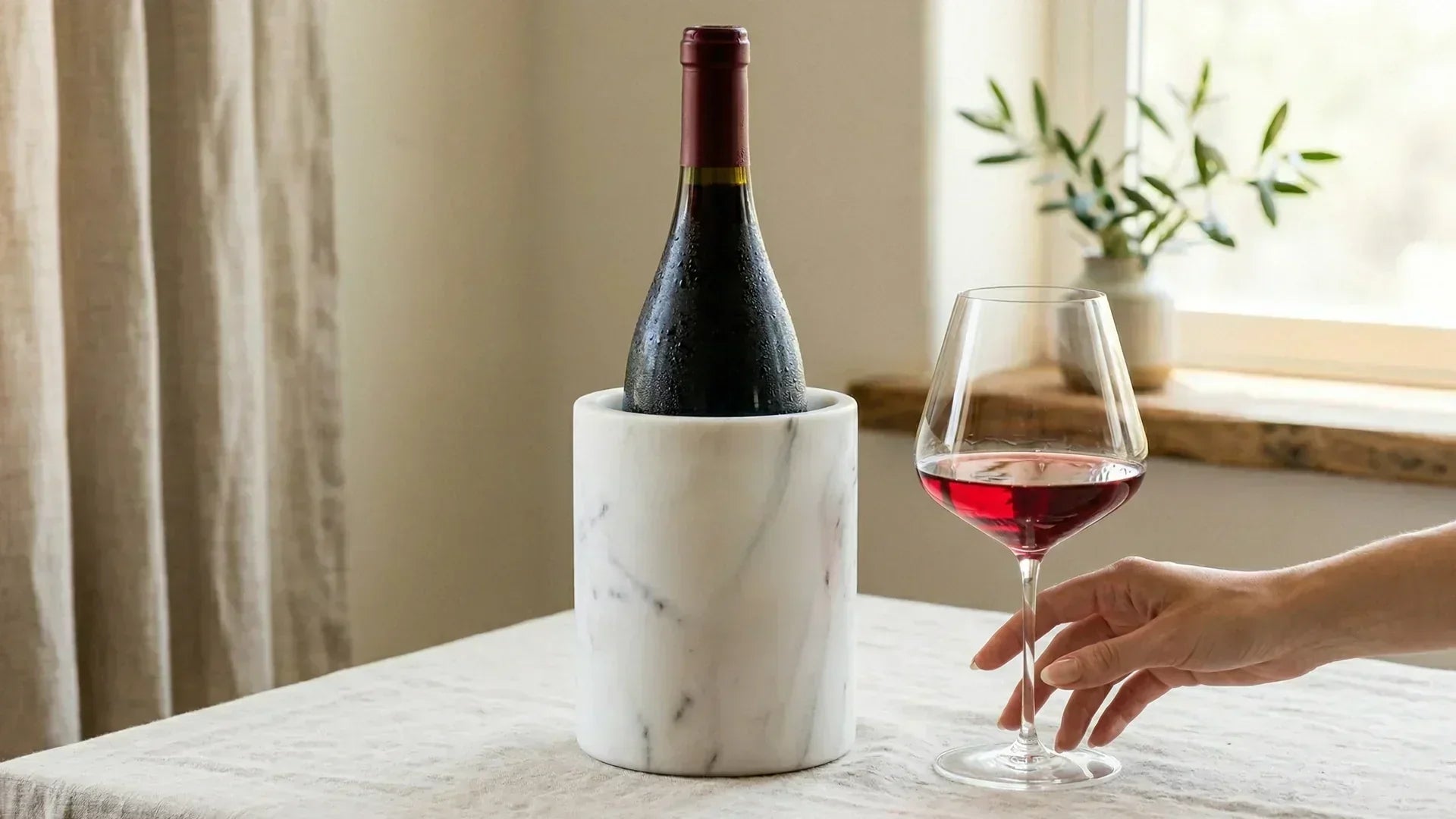 Chilled red wine bottle in marble cooler with glass of light red wine on table by window