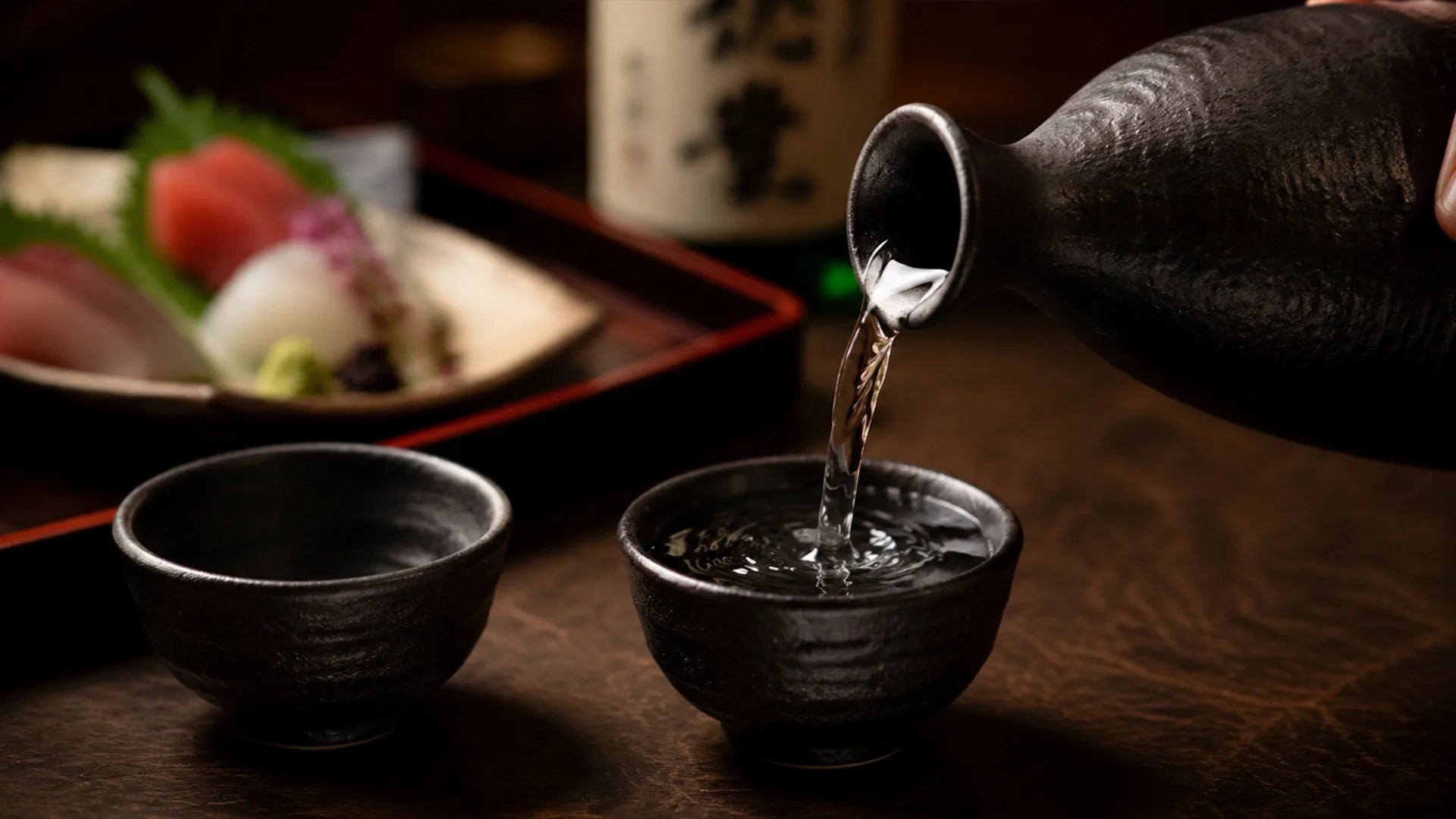 Close-up of clear sake pouring from a dark ceramic tokkuri pitcher into a matching ochoko cup on a rustic wooden table, with fresh tuna and white fish sashimi elegantly plated in the background.