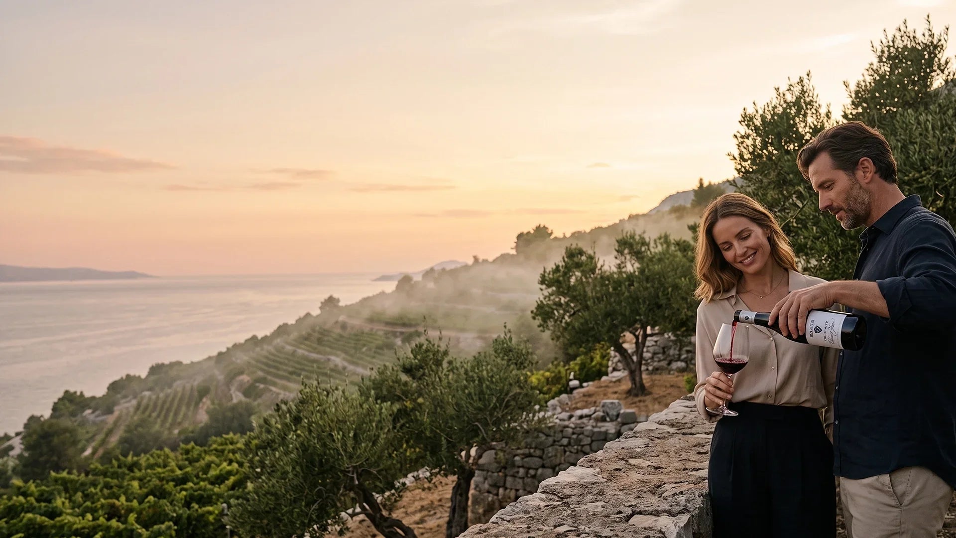A man pouring red wine into a glass for a woman leaning against a stone wall, with a backdrop of terraced vineyards along a misty coastline and sea at sunset.