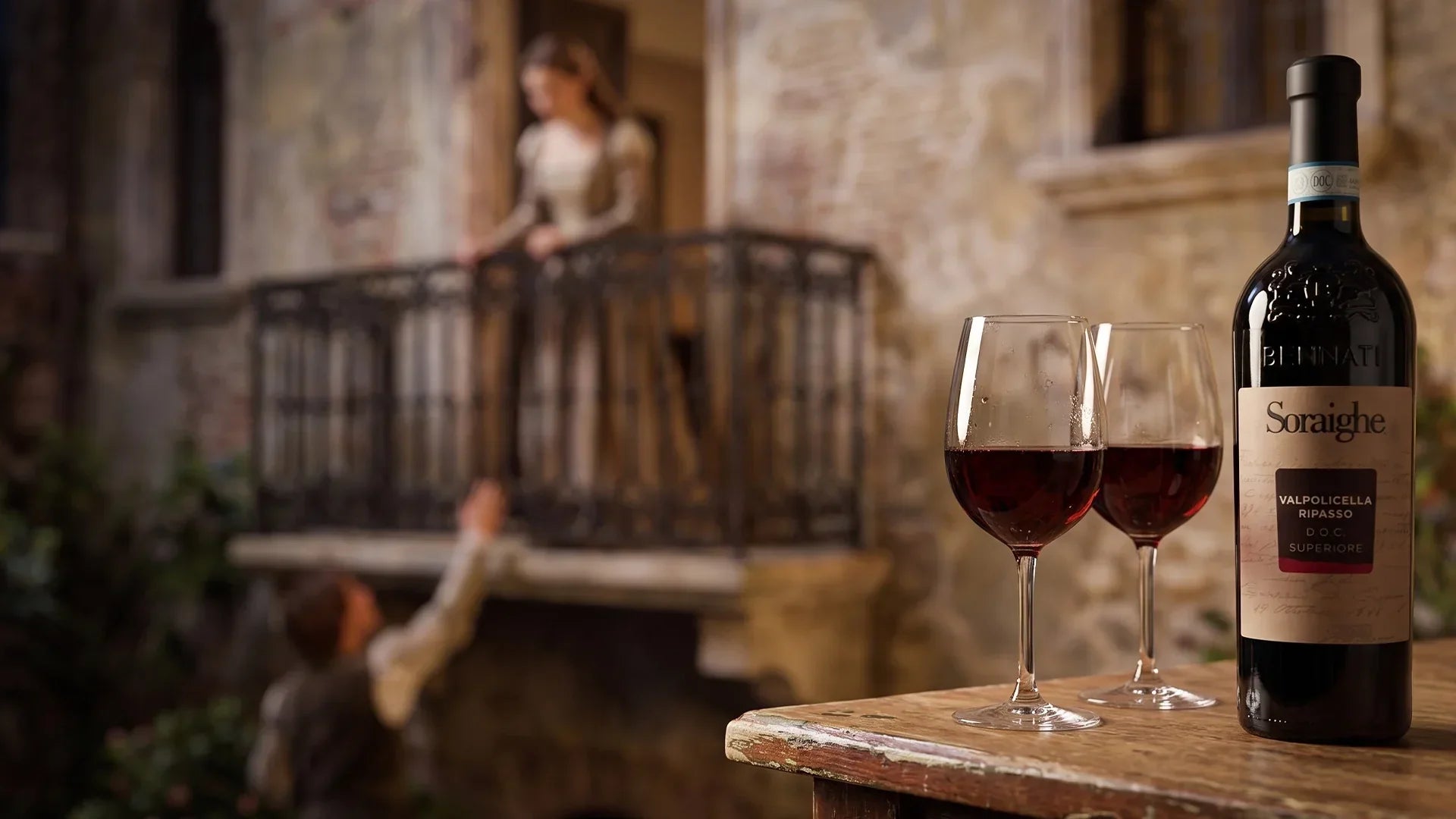 A bottle of Bennati Soraighe Valpolicella Ripasso wine and two filled wine glasses on a wooden table, with a couple re-enacting a romantic balcony scene in the blurred background.