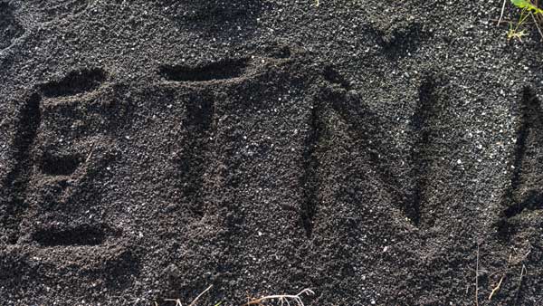 The word "Etna" carved into coarse black volcanic sand