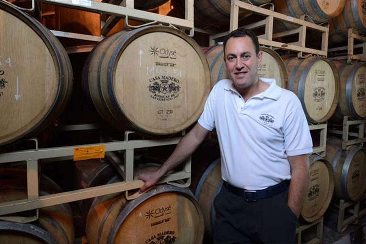 Man in a white polo standing beside stacked oak barrels marked Casa Madero in a cellar