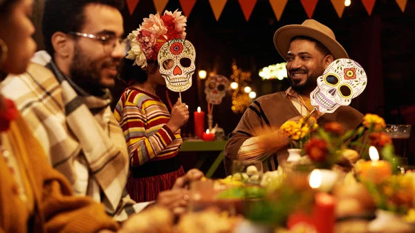 Friends at a candlelit dinner holding decorated sugar skull masks on sticks, marigolds on the table