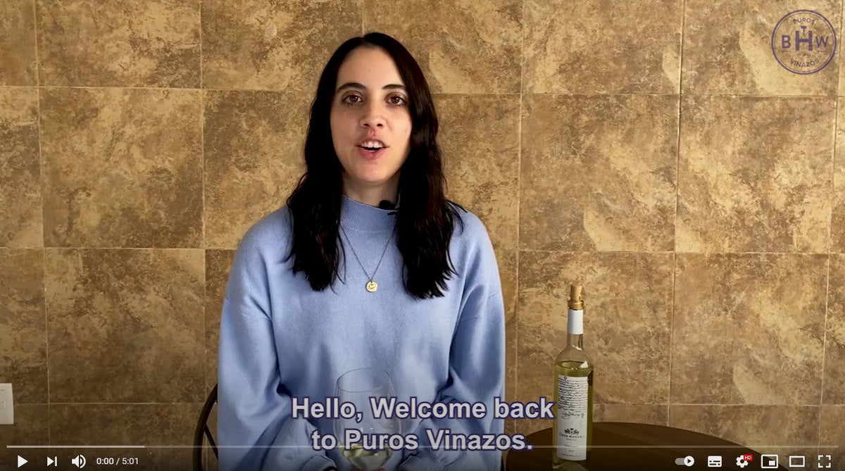 Video still of a woman speaking beside a white wine bottle, captioned 'Hello. Welcome back to Puros Vinazos.'