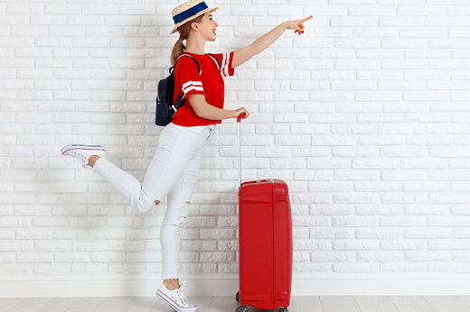 Woman in a straw hat with a backpack pointing ahead, standing beside a red suitcase