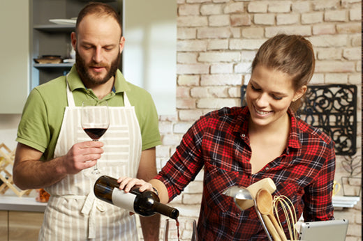 Man in an apron holding a glass of red wine while a woman pours from a bottle in a kitchen
