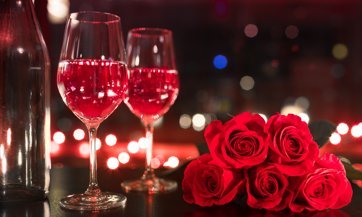 Two glasses of rosé wine beside a bouquet of red roses with bokeh lights behind