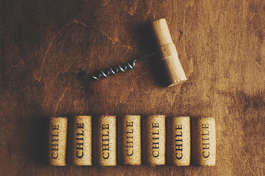 Seven wine corks printed "Chile" laid in a row on wood, with a corkscrew and another cork above