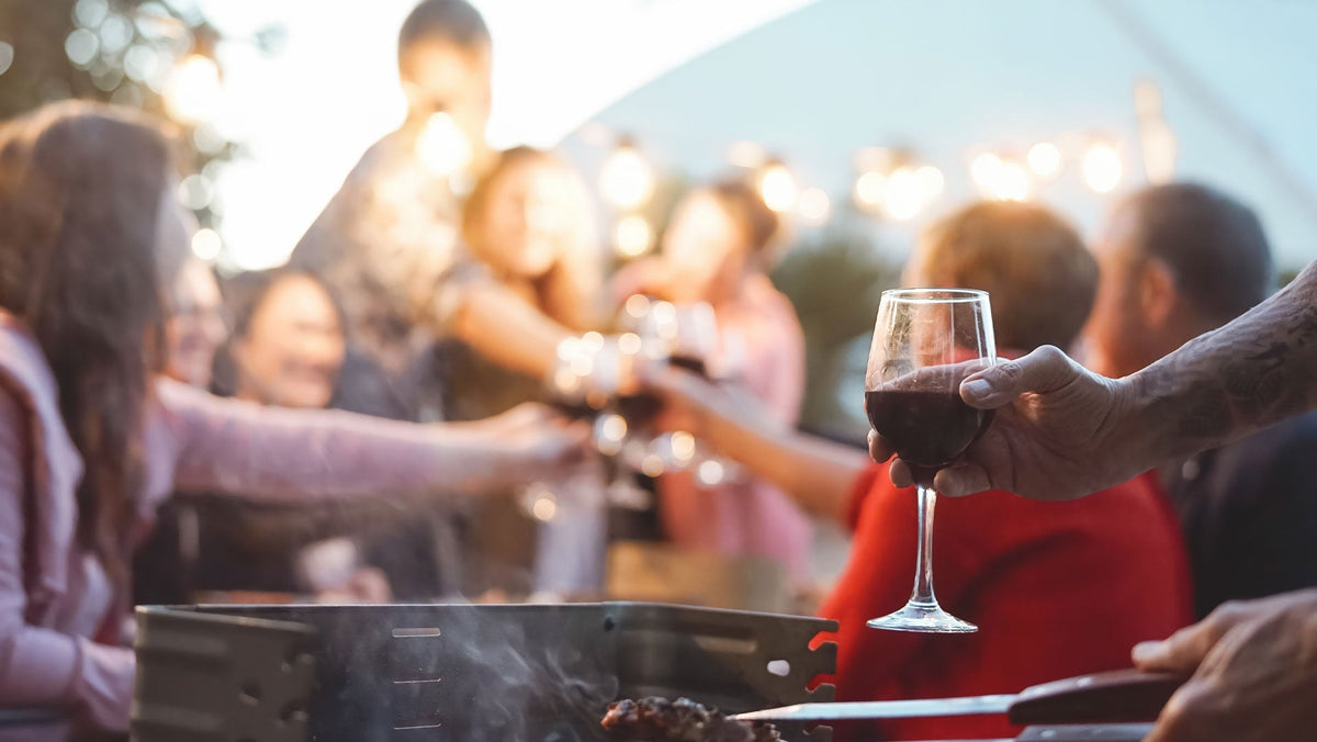 Hand holding a glass of red wine over a smoking grill as a group toasts outdoors at dusk