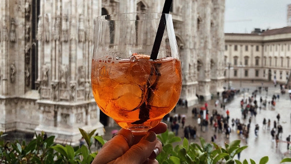 Hand holding an orange spritz cocktail in front of the Milan Duomo on a rainy day