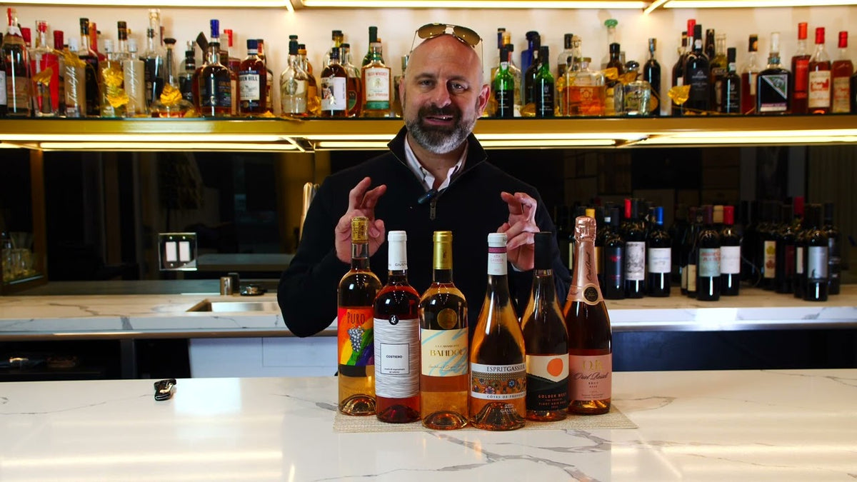 Man behind a marble bar gesturing toward a row of rose and sparkling wine bottles