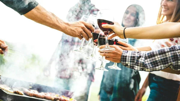 Hands toasting glasses of red wine at an outdoor barbecue with smoke rising from the grill