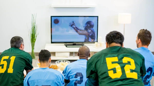 Men in football jerseys numbered 51 and 52 watching a football game on TV, seen from behind