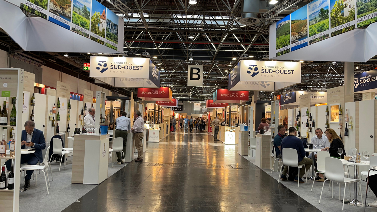 Aisle of exhibitor stands in a trade fair hall with Sud-Ouest signs and a hanging section B marker
