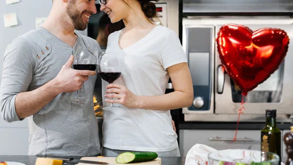 Couple in a kitchen holding glasses of red wine, red heart balloon behind them