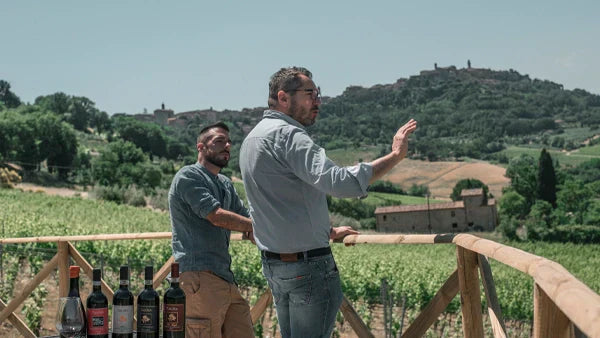 Two men on a vineyard terrace with wine bottles on the rail, gesturing toward a hilltop town