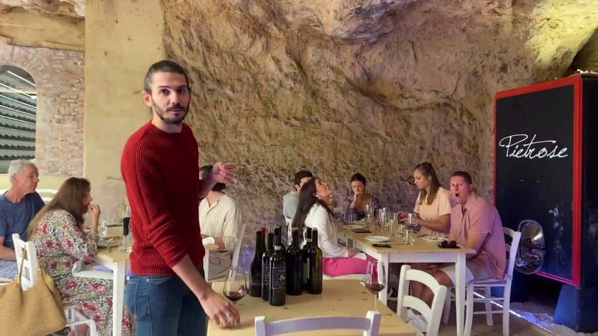 Man standing in a cave-walled tasting room with seated guests and red wine bottles on the tables