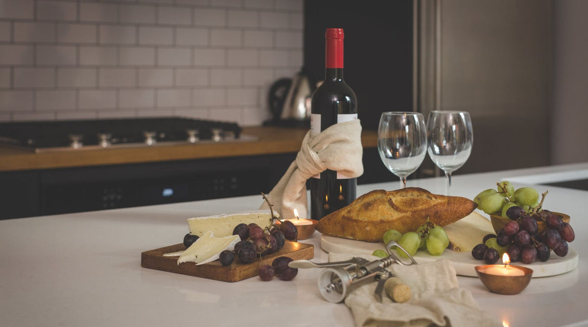 Bottle of red wine, two empty glasses, a cheese board, bread and grapes on a kitchen counter