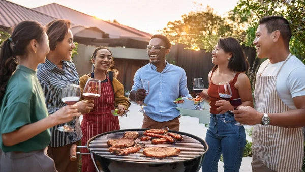 Friends holding wine glasses around a barbecue grill of ribs at sunset