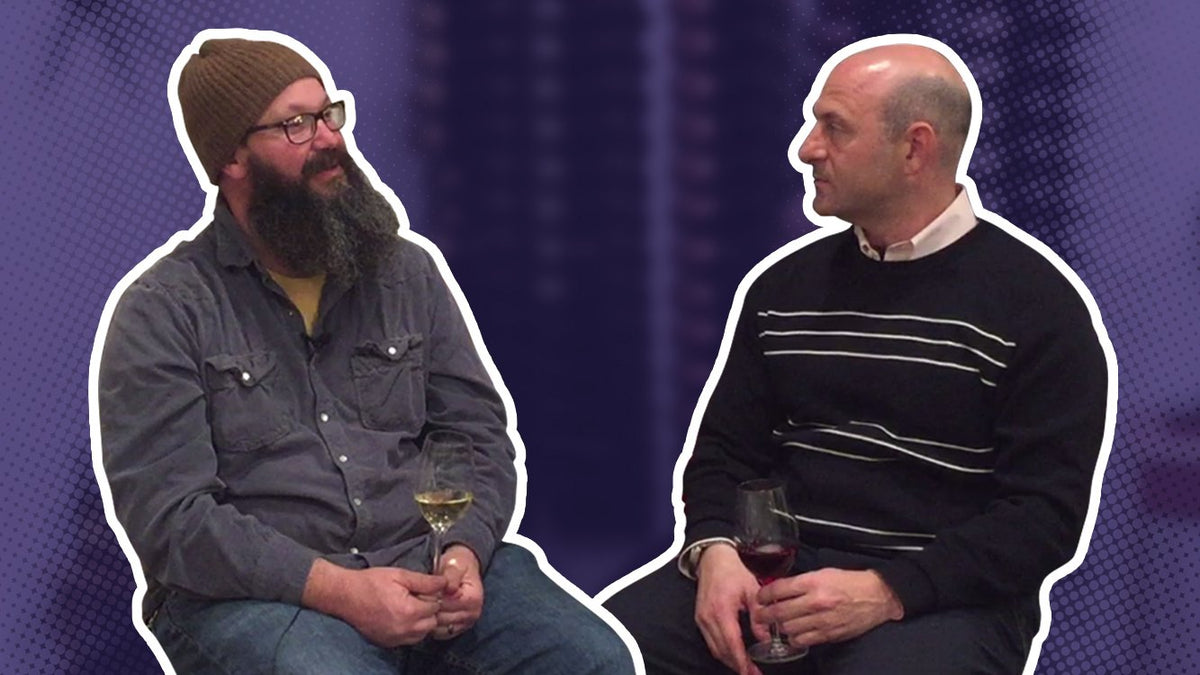 Two men seated holding glasses of white wine during a filmed conversation