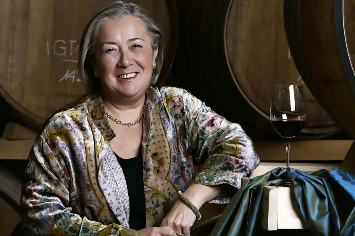 Smiling woman in a floral jacket seated in front of large oak casks with a glass of red wine