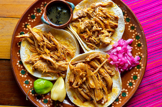 Three cochinita pibil tacos on a clay plate with pickled red onion, limes and a bowl of salsa