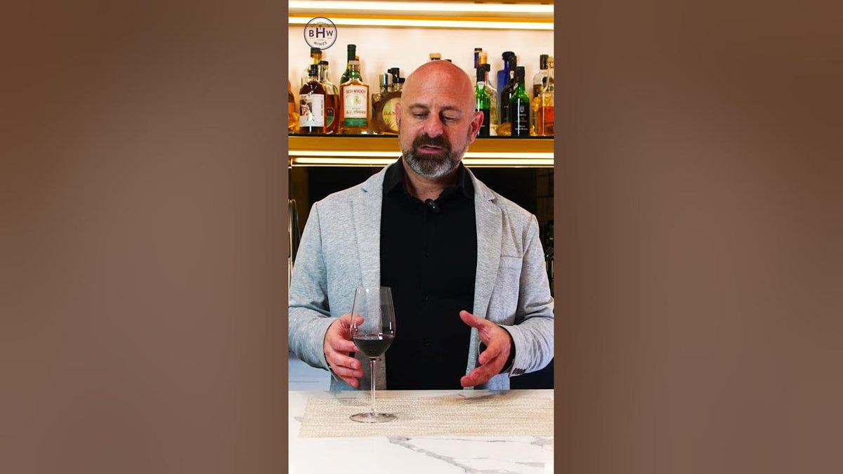 Man in a grey blazer holding a glass of red wine at a marble bar, bottles on shelves behind