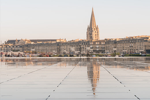 Riverfront buildings and a church spire reflected in a shallow water mirror, Bordeaux