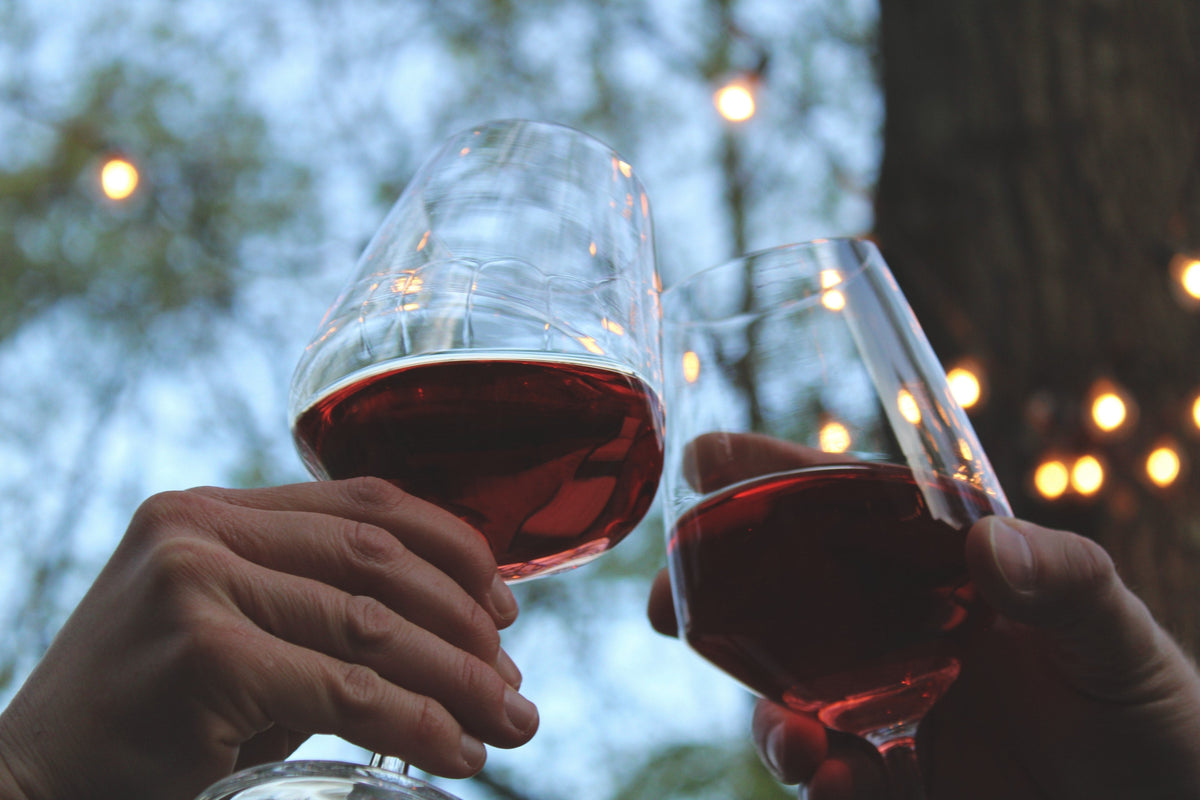 Two hands clinking glasses of red wine outdoors under string lights at dusk