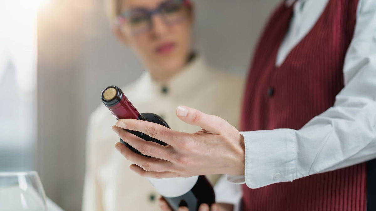 Server in a burgundy vest presenting a bottle of red wine to a woman in white