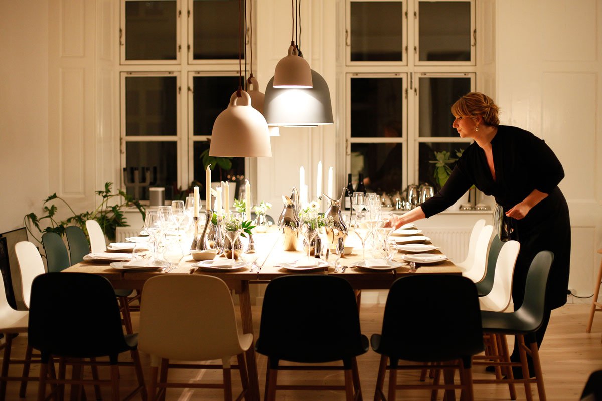 Woman setting a long dining table with candles, wine glasses and place settings under pendant lamps