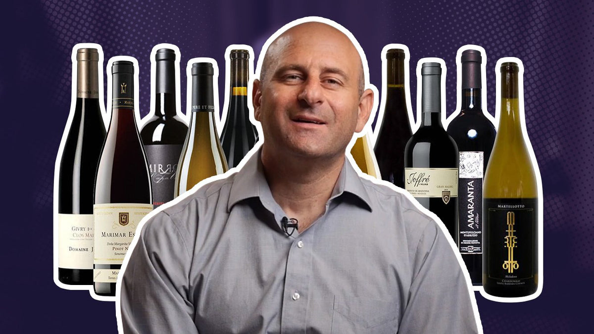 Man in a grey shirt surrounded by ten bottles of red and white wine