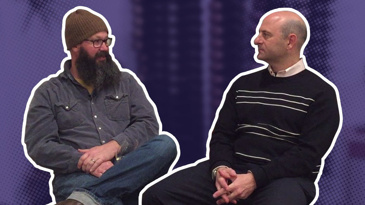 Bearded man in a beanie seated in conversation with a man in a striped sweater