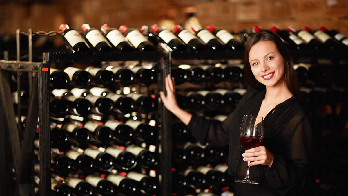 Woman holding a glass of red wine beside a full wine rack in a cellar