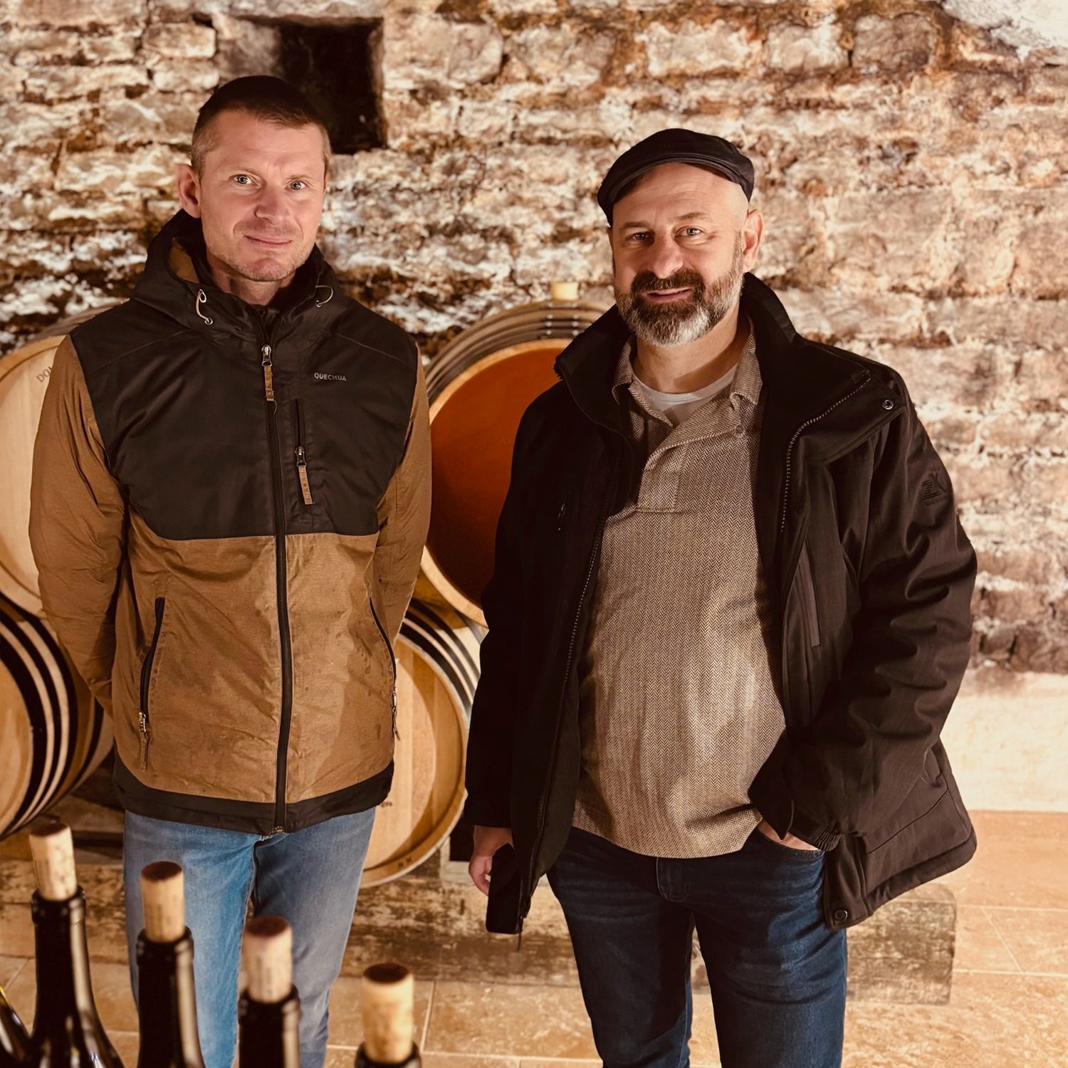 Greg Martellotto and Alexis Guillon from Domaine Guillon & Fils winery in Gevrey-Chambertin, sharing insights on the winemaking process amidst barrels and bottles of wine.