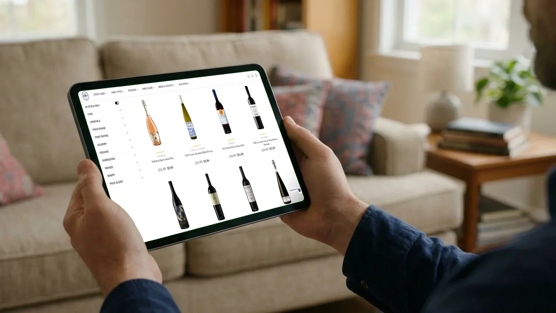 Person browsing an online wine shop on a tablet at home, comparing bottles and prices