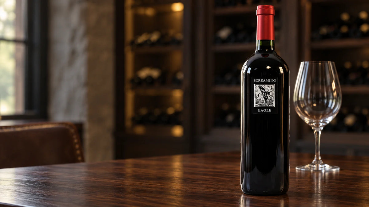 A bottle of Screaming Eagle red wine with a red foil cap standing next to an empty wine glass on a dark wooden table in a cellar setting.   