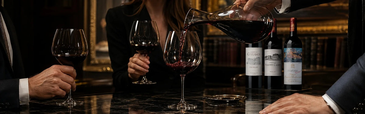 A person pouring red wine from a decanter into two wine glasses on a dark bar counter with Pauillac wine bottles and parts of two people visible in a dimly lit indoor setting.