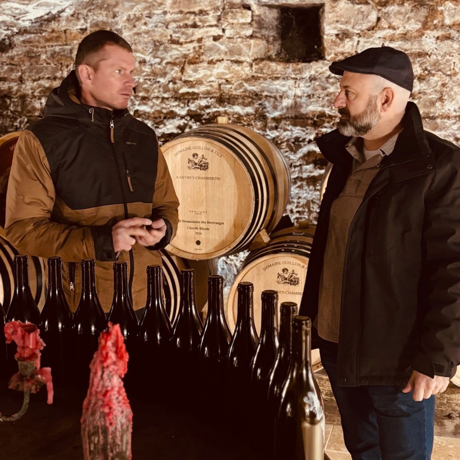 Greg Martellotto and Alexis Guillon from Domaine Guillon & Fils winery in Gevrey-Chambertin, sharing insights on the winemaking process amidst barrels and bottles of wine.