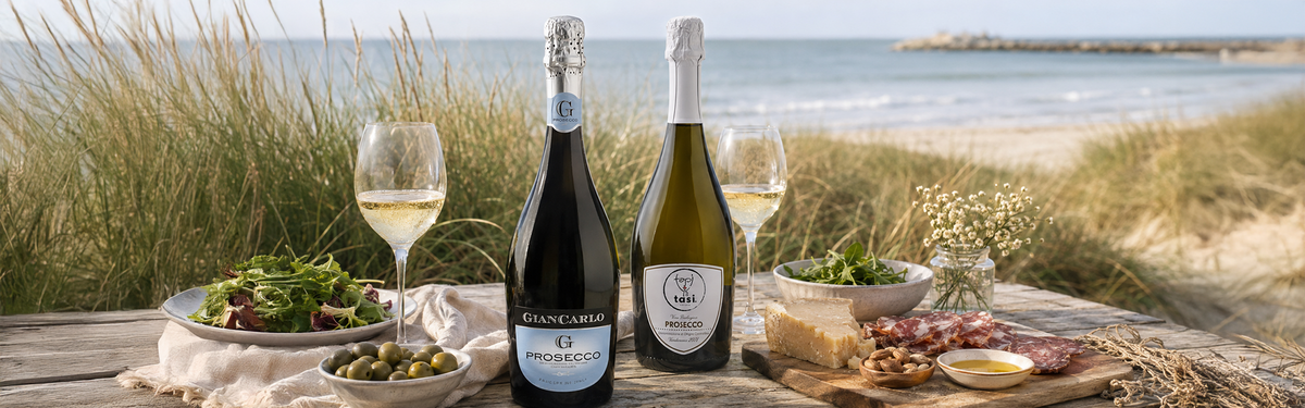 Two bottles of Prosecco with filled wine glasses, salad, and a charcuterie platter on a beachside wooden table.